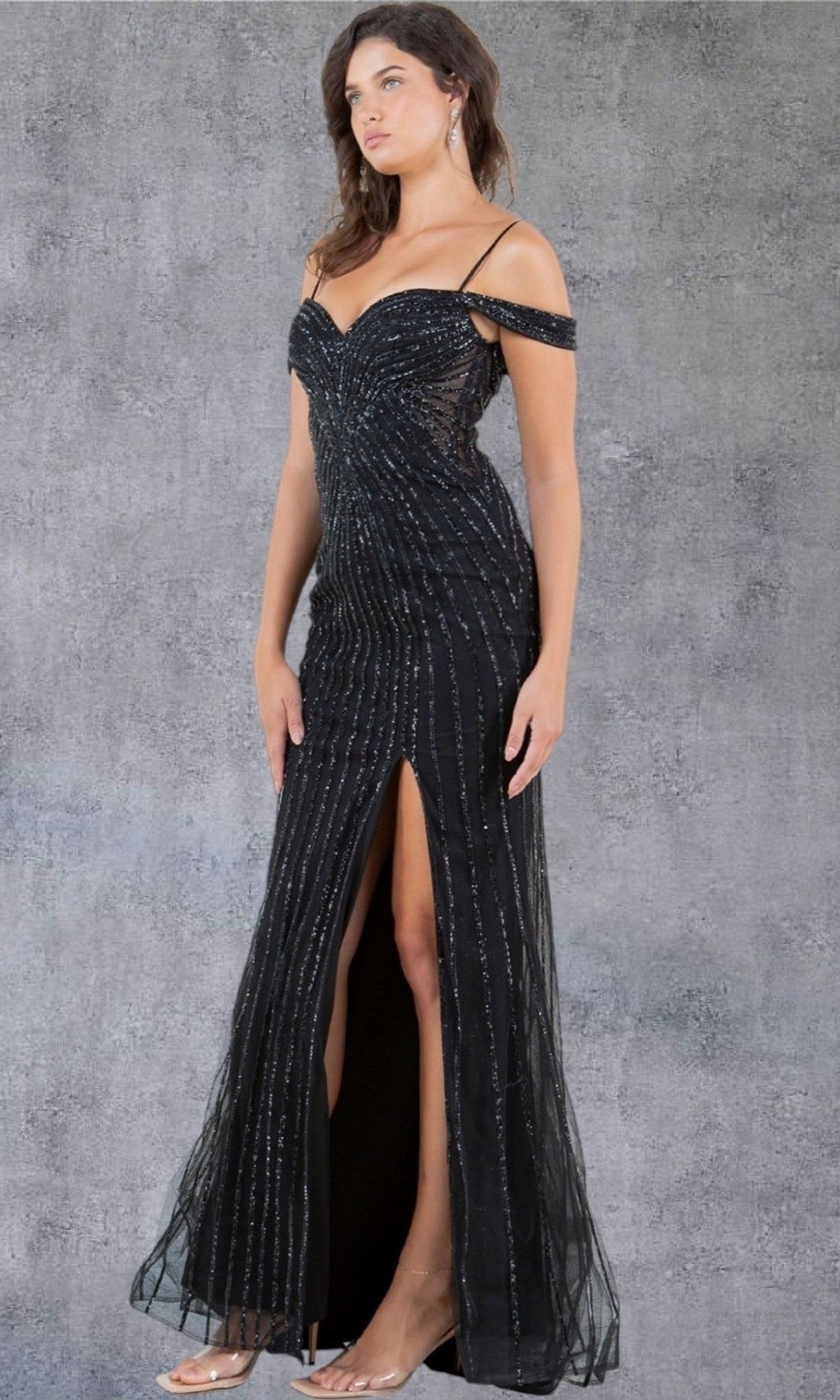 A woman wearing the Cinderella Couture 8269J long prom dress in black, featuring thin straps, silver vertical stripes, an off-shoulder neckline, and a floor-length silhouette with a front thigh-high slit, stands against a gray textured background.